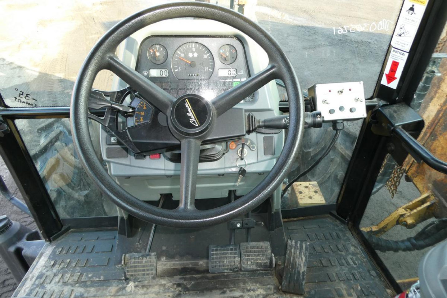 Industrial tractor cabin view with steering wheel and dashboard, pedals visible, operator controls on left and right side, dusty cockpit interior.