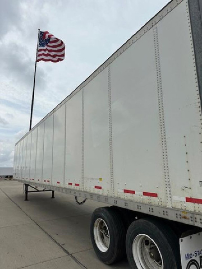 Trailer side view showing white Wabash van trailer with US flag in background; riveted side panels, tandem axles, reflective red/white marker strips.