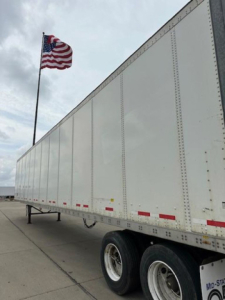 Trailer side view showing white Wabash van trailer with US flag in background; riveted side panels, tandem axles, reflective red/white marker strips.