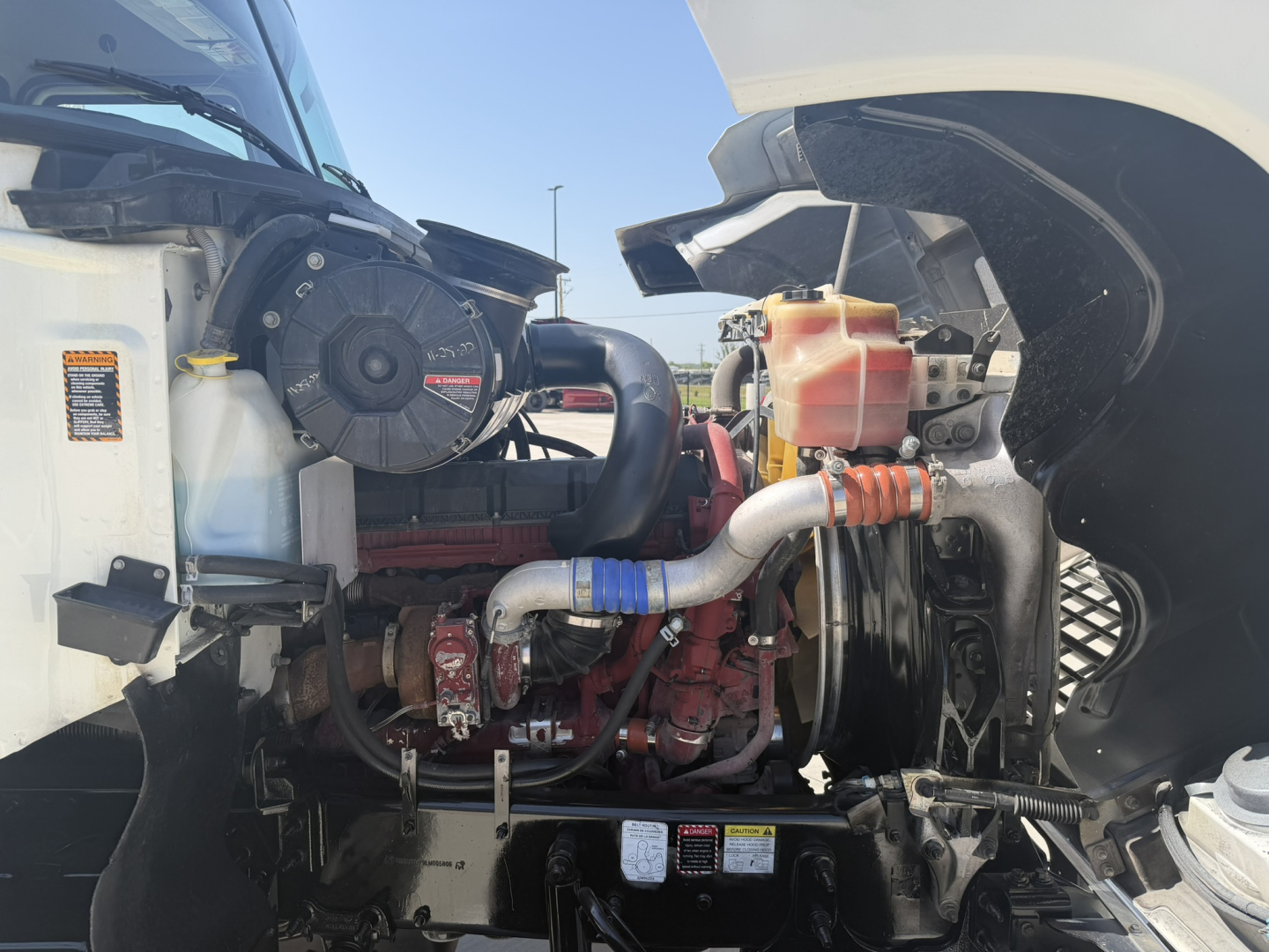 Mack PI64T engine bay showing coolant reservoir, air intake, radiator hose clamps, and turbocharged engine visible under open hood.