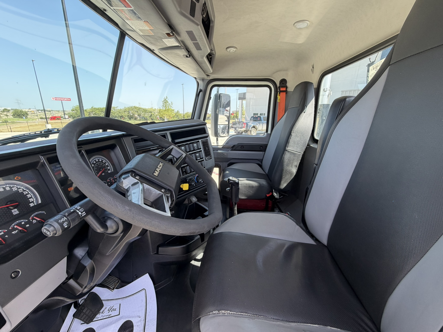 Truck cab interior showing Mack PI64T dashboard, steering wheel, and front seats with light gray/black upholstery; sun visor; windshield; gear shifter visible; instruction paper on floorboard.