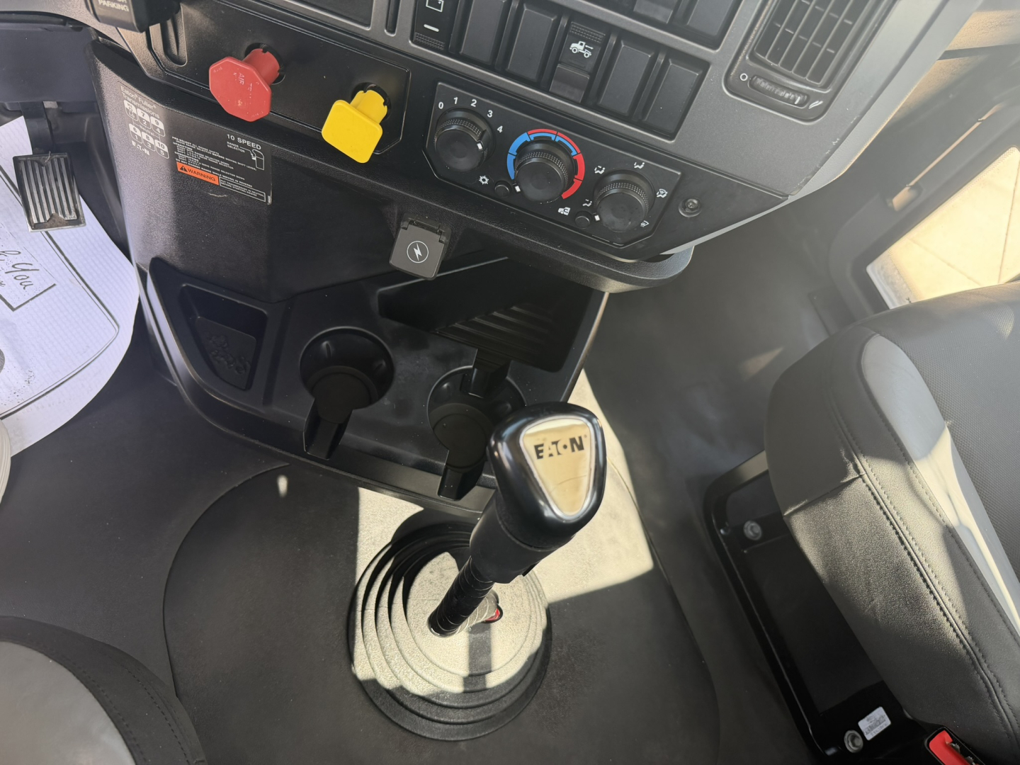 Mack PI64T interior gear shifter and dashboard with red and yellow controls, manual gear lever in foreground, circular climate controls, and driver seat visible.