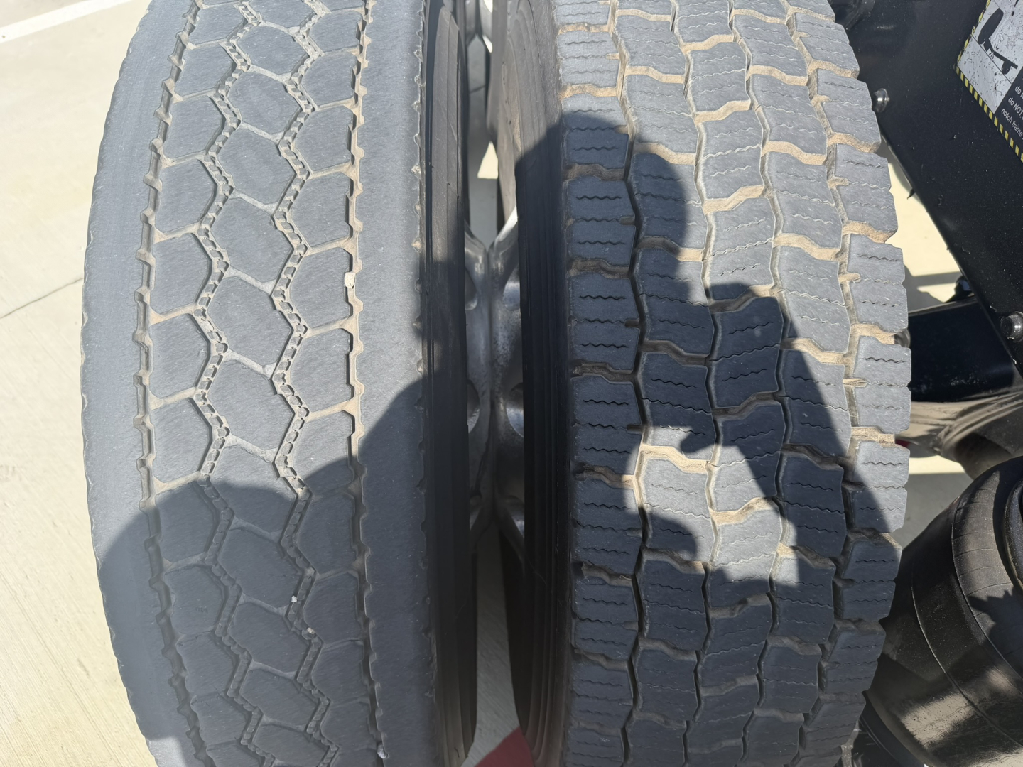 Rear dual Mack PI64T truck tires with tread patterns visible; left tire smoother, right tire deeper tread, in a daycab truck configuration.