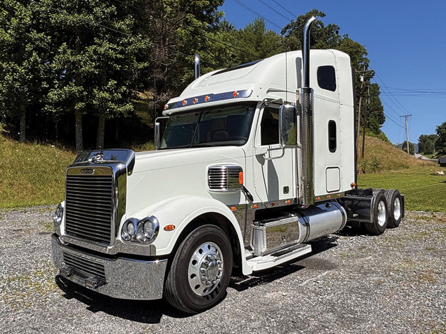 Truck cab showing Freightliner Coronado sleeper with chrome bumper and dual rear axles, white exterior, side view mirrors, and vertical exhaust stack.