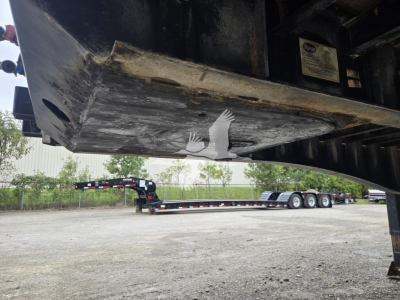 Underneath a steel flatbed trailer showing the underside and a label plate; the ground and another trailer in the background.