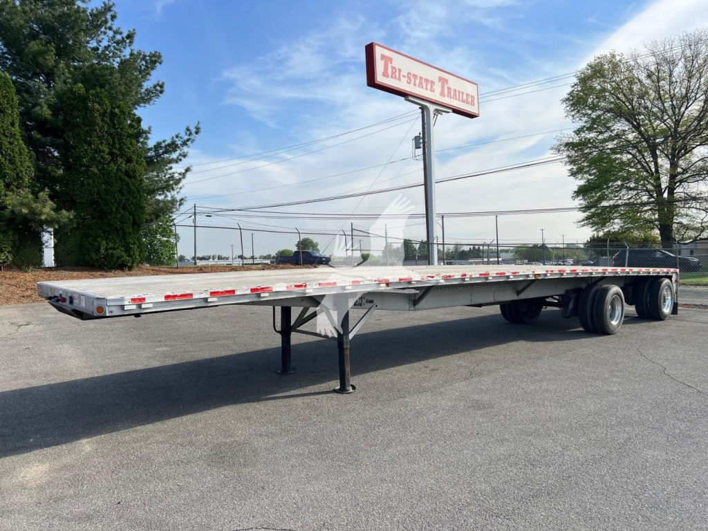 Flatbed trailer showing extended platform, landing legs deployed, tandem axle rear, reflective marker lights along edge, Tri-State Trailers sign in background.