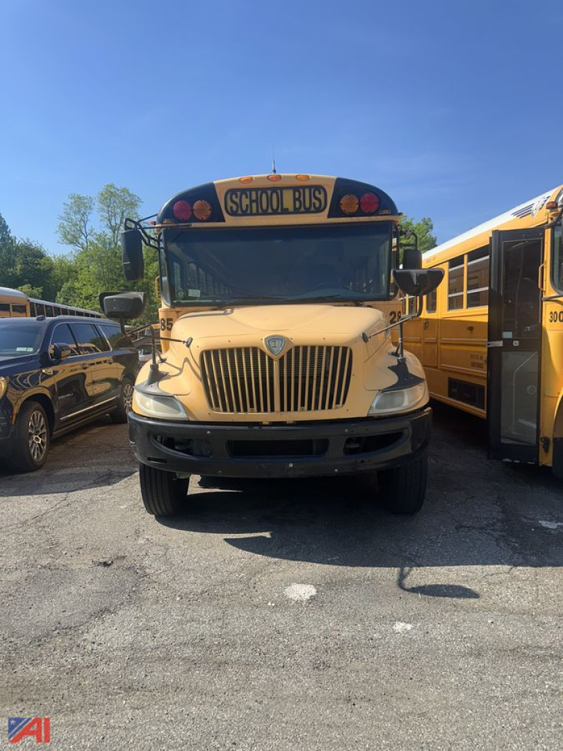 School bus front view showing CE Series logo emblem, beige body, large grille, and 'SCHOOL BUS' sign on top, with other yellow bus on the right.