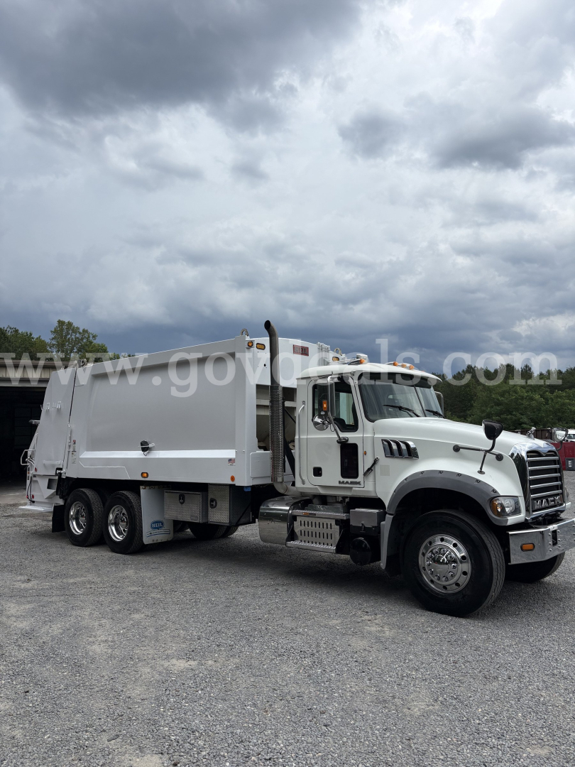 Mack GU713 garbage truck in-service with white cab and refuse body, three axles, side view. Visible text on image: none legible; generic truck image.
