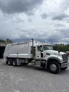 Mack GU713 garbage truck in-service with white cab and refuse body, three axles, side view. Visible text on image: none legible; generic truck image.