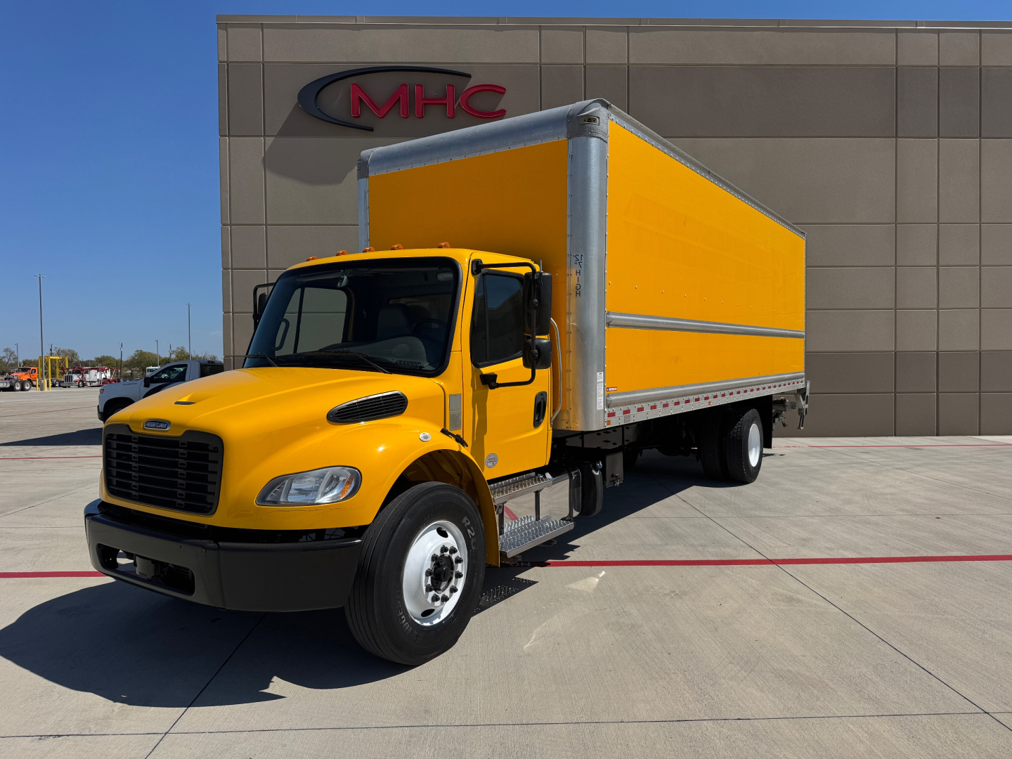 Box truck with yellow cab and yellow box body in front of a building labeled MHC.