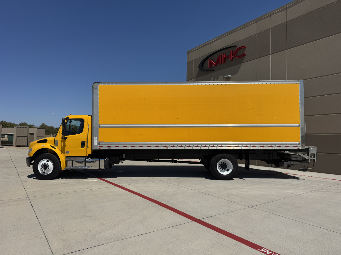 Truck side view of a yellow Freightliner M2-106 box truck, long cargo box, at a dealership lot, with a building in background.