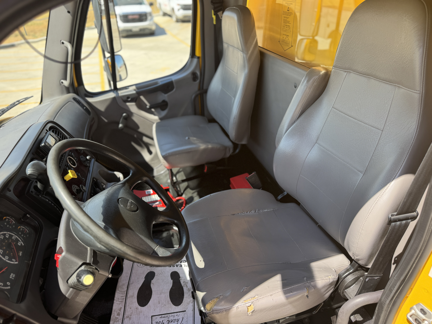 Cab interior of a 2022 Freightliner M2-106 with front driver seat and passenger seat, steering wheel, dashboard, and gear shifter visible.