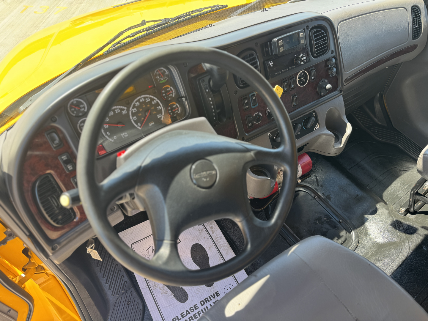 Cab interior of a yellow Freightliner M2-106 showing steering wheel, dashboard, gauges, radio, and center console.