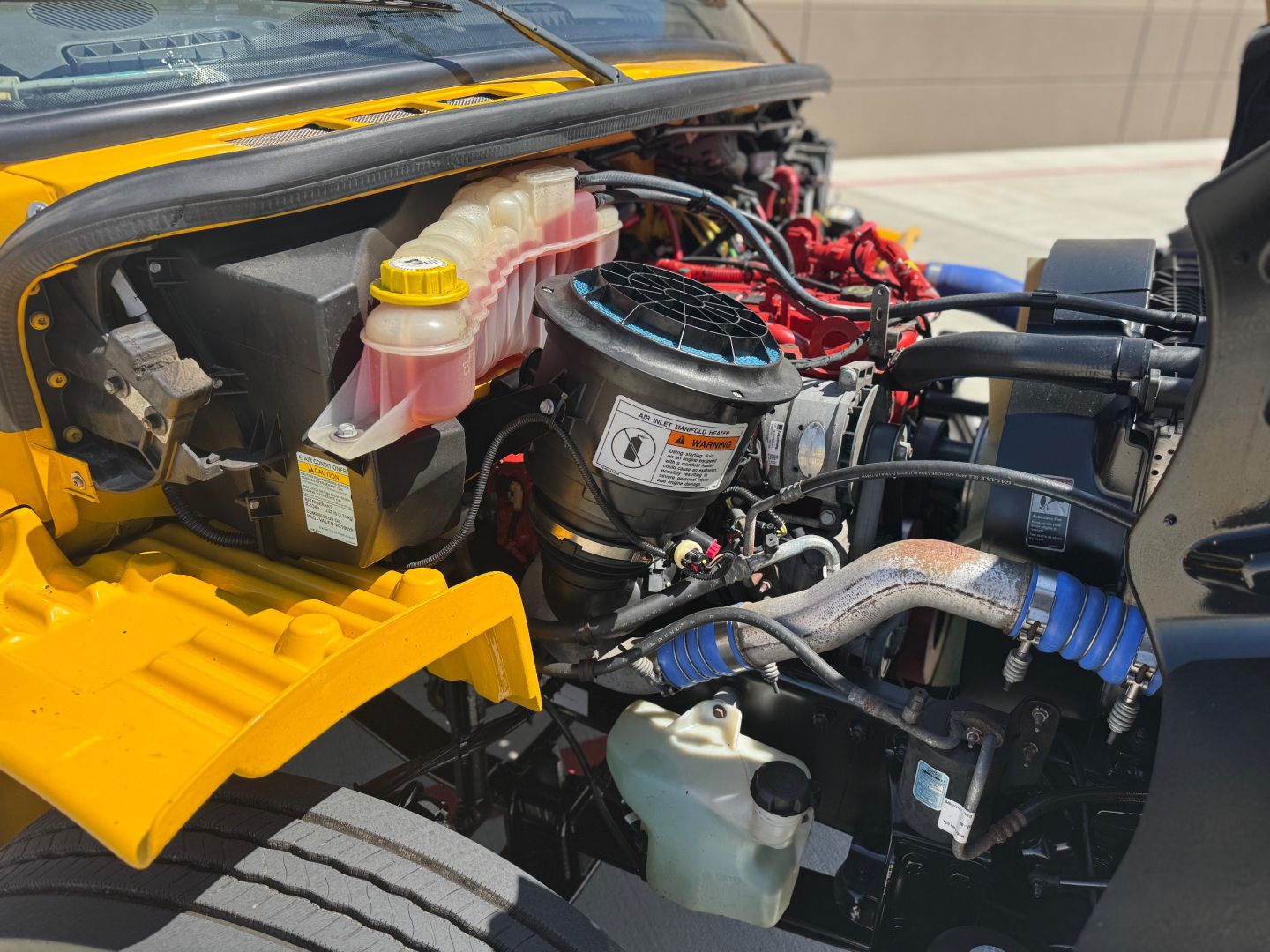 Freightliner M2-106 engine bay showing coolant reservoir, pink radiator fluid reservoir, yellow cap, black air intake housing, and various hoses and components.