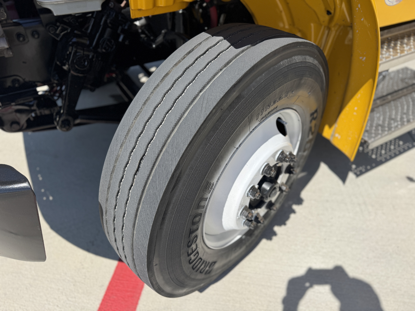 Freightliner M2-106 front tire and wheel assembly in view showing Bridgestone tire and multi-lug hub, yellow bodywork nearby.