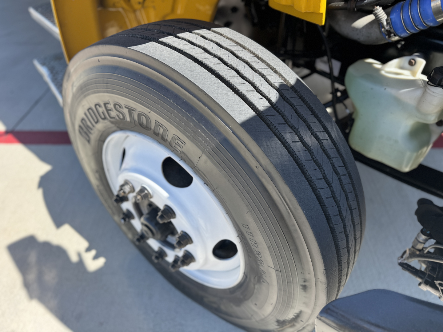 Bridgestone tire on Freightliner M2-106 front wheel well with brake drum and wheel hub, visible cab area components and a coolant reservoir in the background.