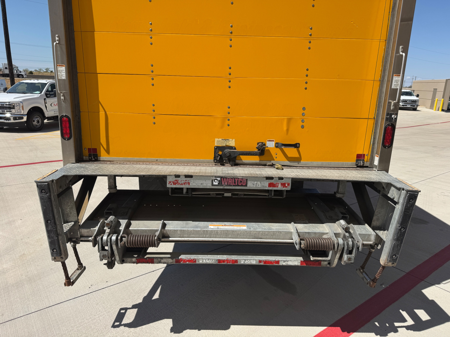 Back of a Freightliner M2-106 box truck showing a hydraulic lift/ram system on the rear with yellow cargo body. Visible stickers/readouts include small labels on the bumper and tailgate hardware.