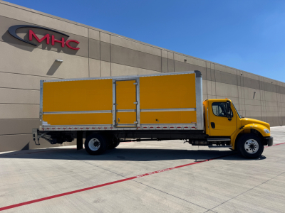 Yellow Freightliner M2-106 box truck parked outside a building with MHC sign; side view shows yellow cargo box and cab, inventory lot setting.