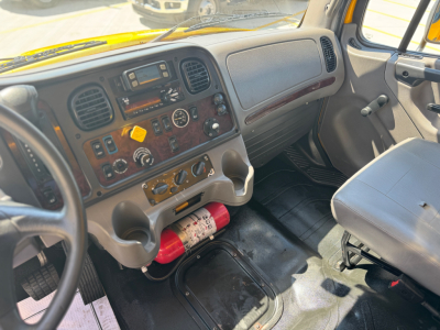 Cab interior of a Freightliner M2-106 showing dash with gauges, radio, climate controls, steering wheel, and a red fire extinguisher beneath the dashboard.
