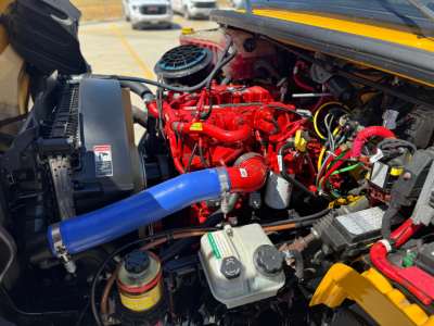 Truck engine bay of a 2022 Freightliner M2-106 showing a red engine, blue intercooler hose, white coolant reservoir, and various cables and hoses.