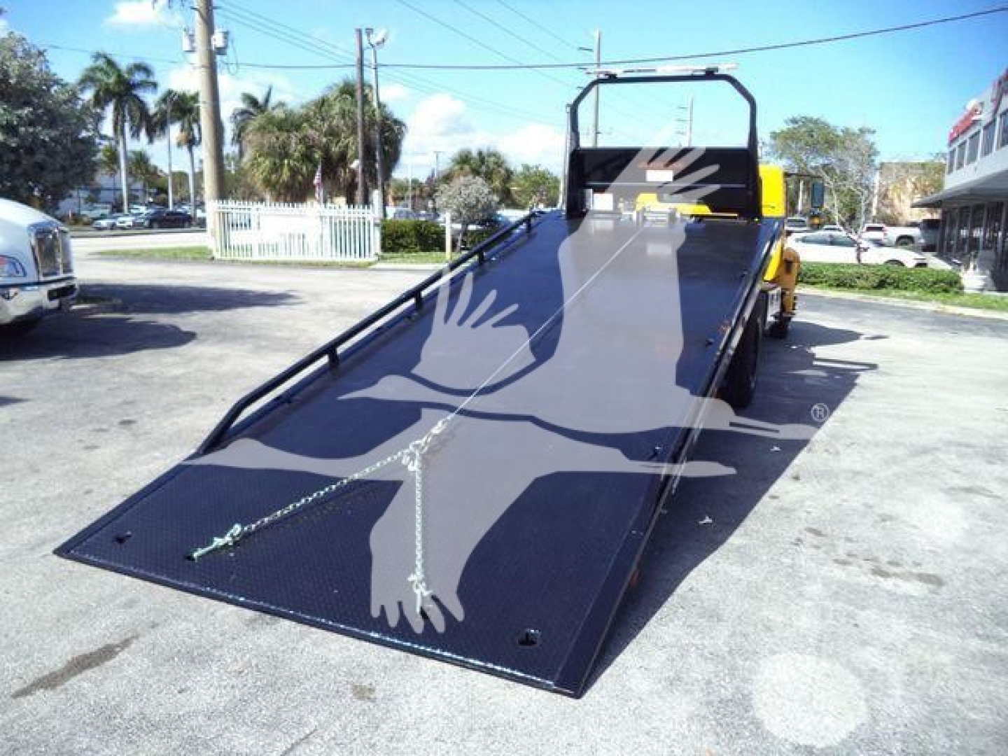 Flatbed/roll-back tow truck with a lowered ramp in a parking lot, black ramp surface, and safety rails.