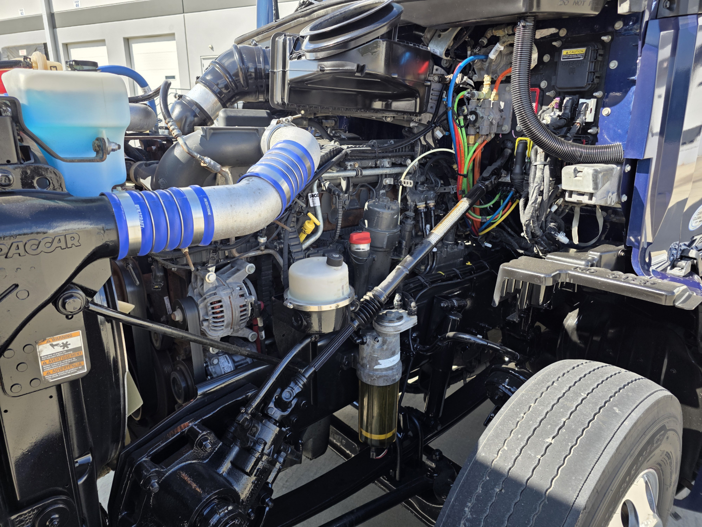 Kenworth T680 engine bay showing blue intake hose, fluid reservoirs, wiring, and engine components.