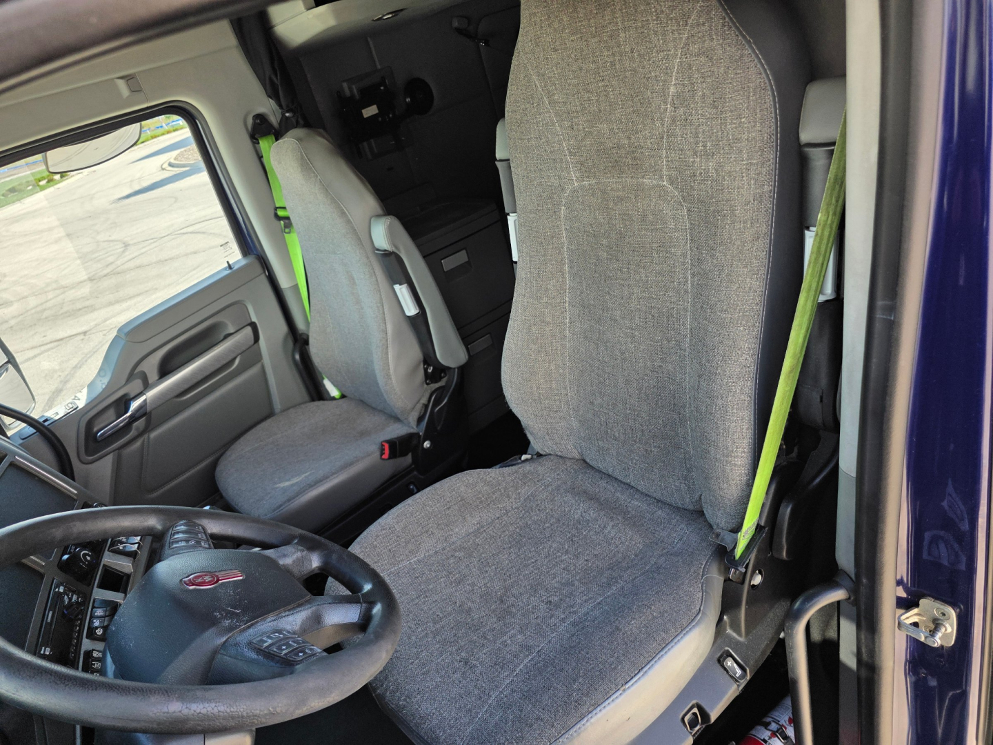 Kenworth T680 interior cab with driver's seat and steering wheel visible; gray fabric seat, door panel, seat belt, and center console area.