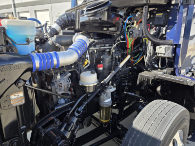 Kenworth T680 engine bay showing blue intake hose, fluid reservoirs, wiring, and engine components.