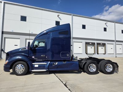 Kenworth T680 truck side profile, blue cab with sleeper, three rear axles, chrome wheels, dealer lot background.