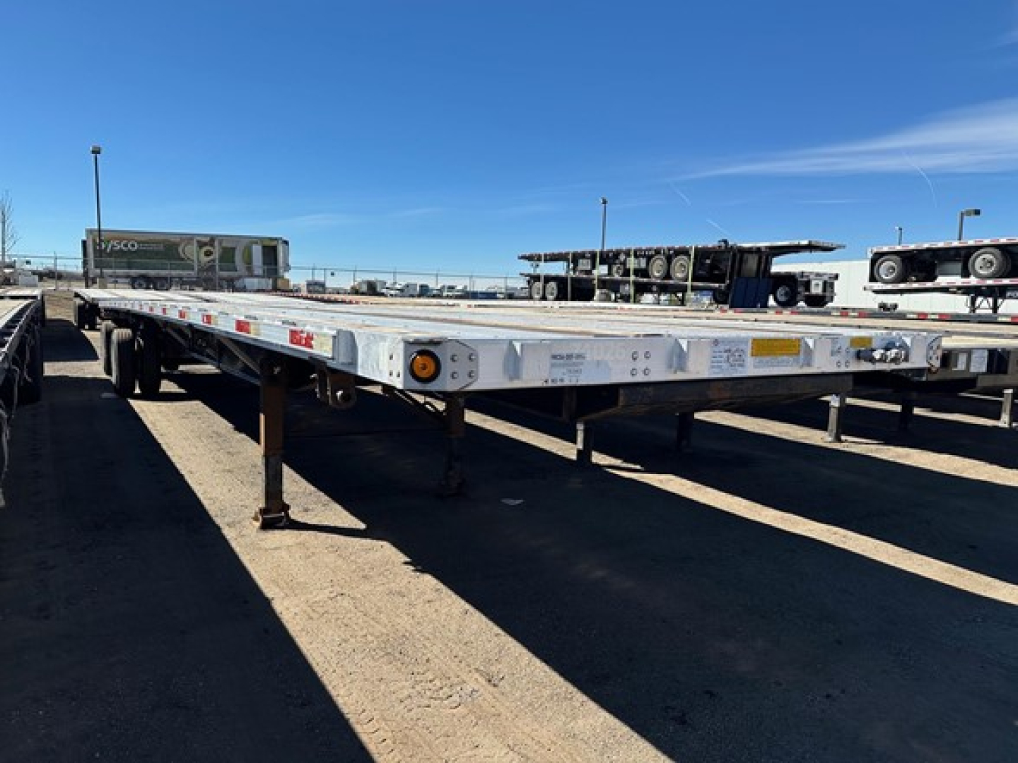 Flatbed trailer with a low-profile deck, side rails, and multiple support jacks visible. Empty gooseneck area; highway trailer lot in background with other trailers and a clear blue sky.