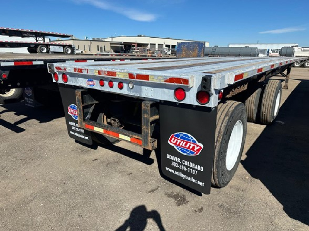 Flatbed trailer showing Utility brand mud flaps and dual rear axles with reflective red/white tape along the sides and back; metal deck with wear; Denver, Colorado stickers on mud flaps.