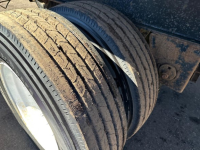 Flatbed trailer wheel twin tires with dirt and wear showing on tread and rim. Part of utility trailer undercarriage visible.
