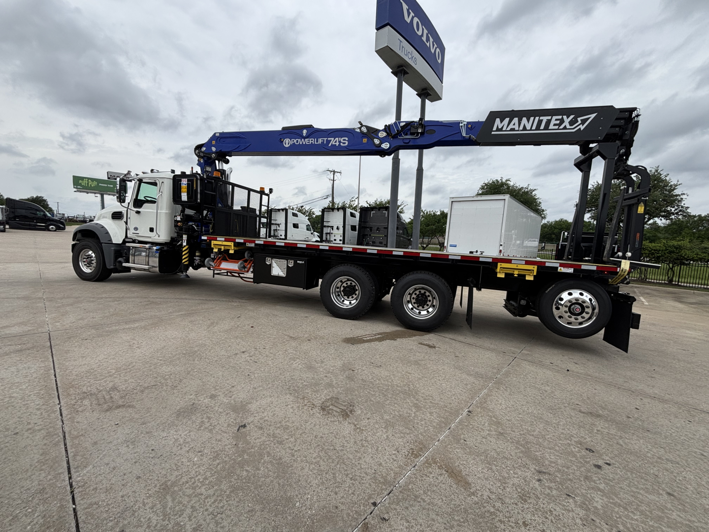 Mack GR64F crane truck with long blue Manitex boom mounted on a white cab and black trailer. Visible text includes MANITEX on the boom, POWERLIFT 7S, VOLVO sign overhead, and various labels along the crane.
