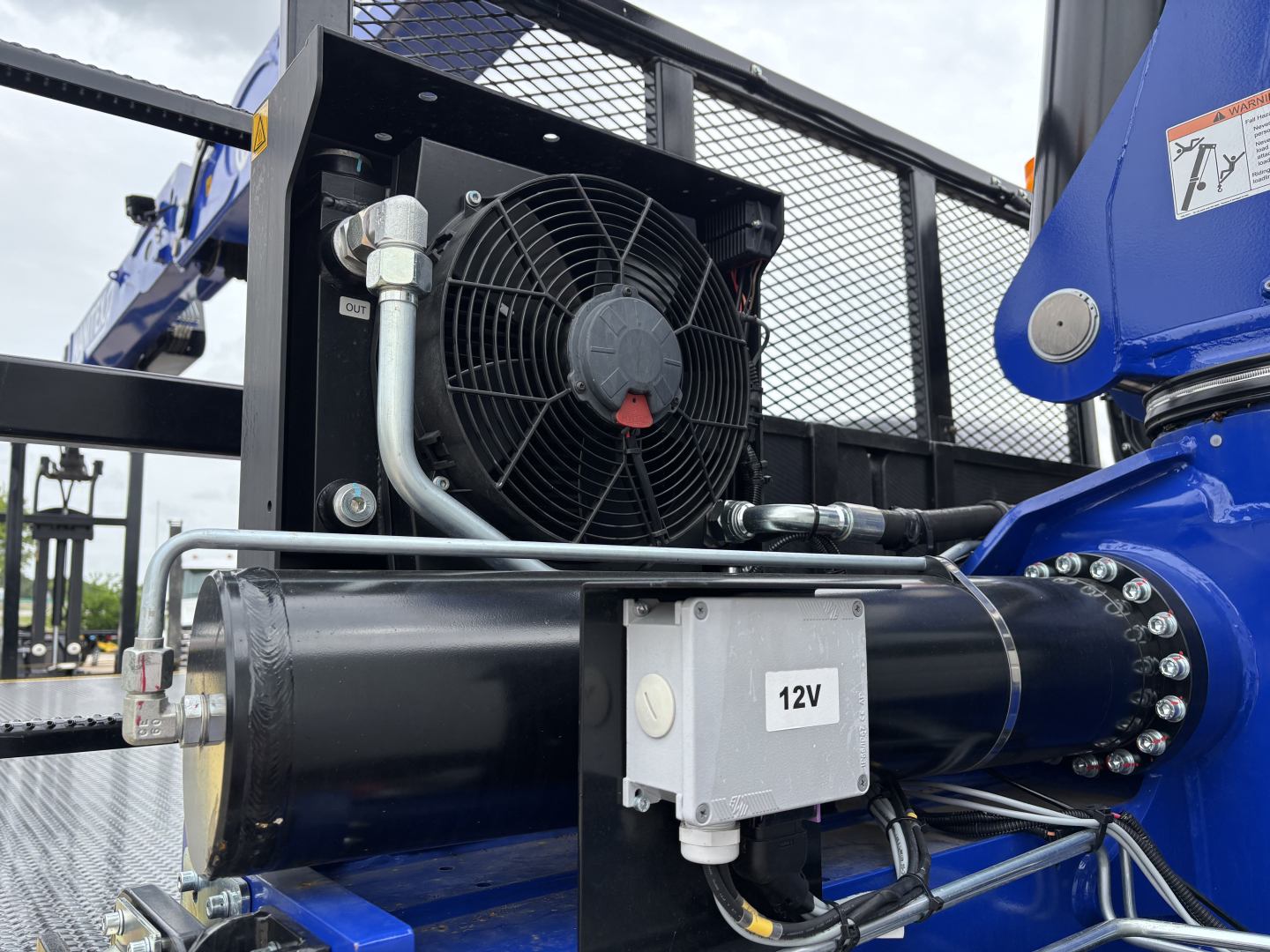 Crane truck engine bay area showing 12V auxiliary control box, radiator fan, hydraulic hose, and blue boom component on Mack GR64F.