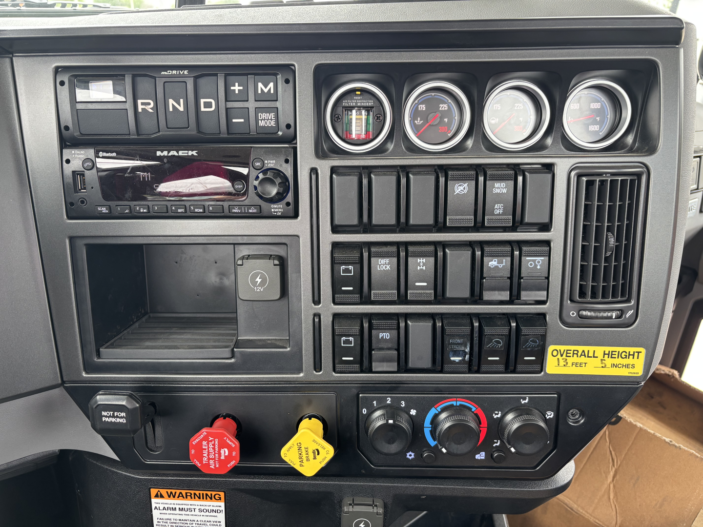 Mack GR64F truck dashboard showing gear selector, gauges, switches, and warning labels; overall height note and control knobs visible.