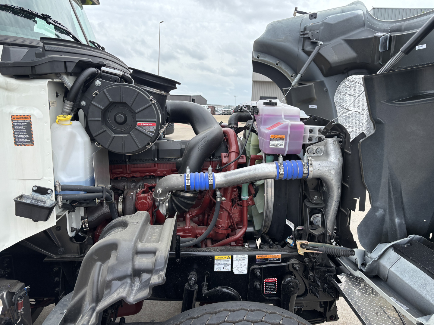 Mack GR64F engine bay with visible turbocharger, cooling reservoirs, hoses, and pink coolant tank. Labeling and components visible from driver side.