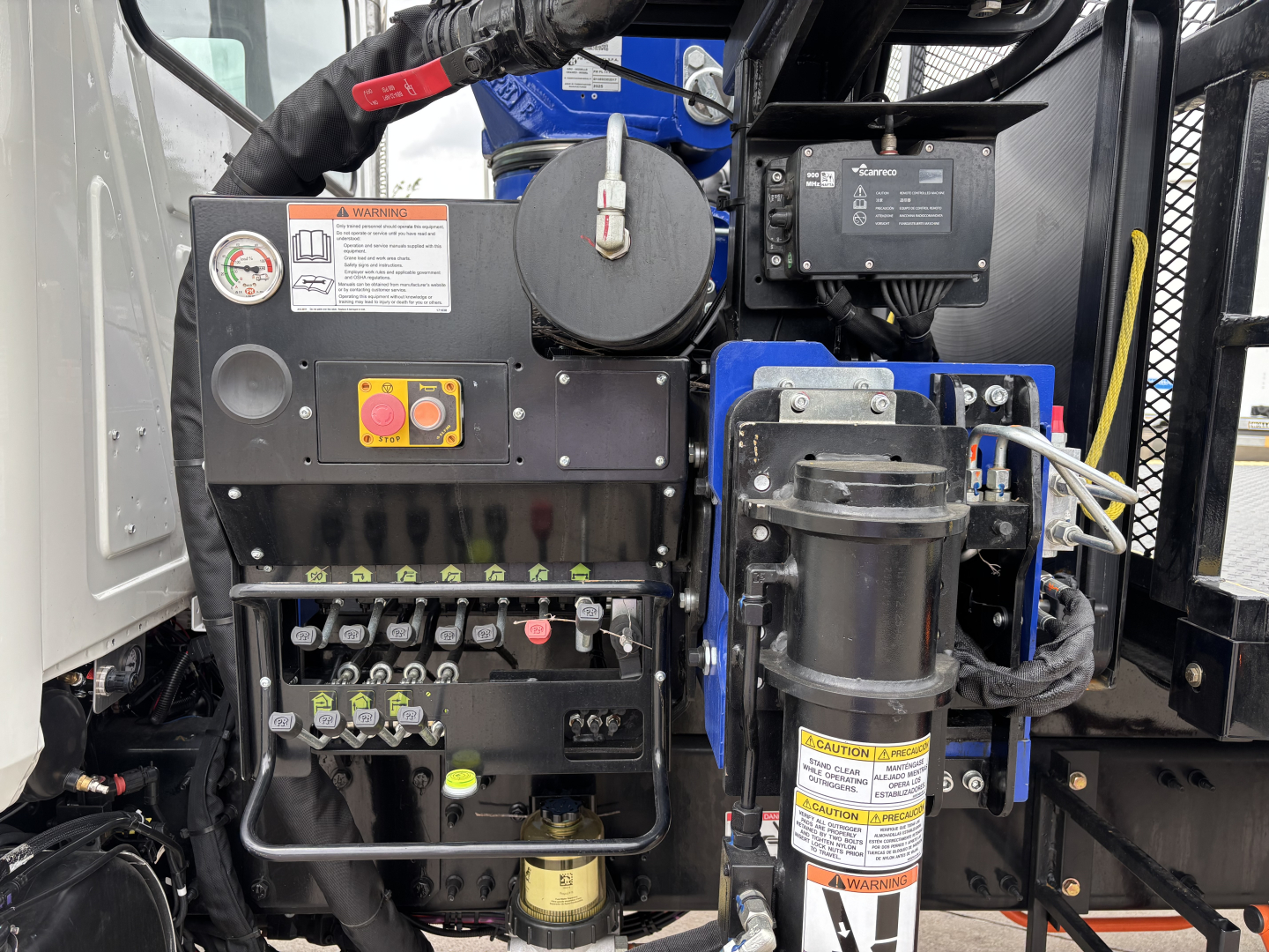 Mack GR64F crane truck engine-bay area showing control valves, hydraulic manifold, warning labels, and cylindrical accumulator. Visible text includes warning placards and control switches.