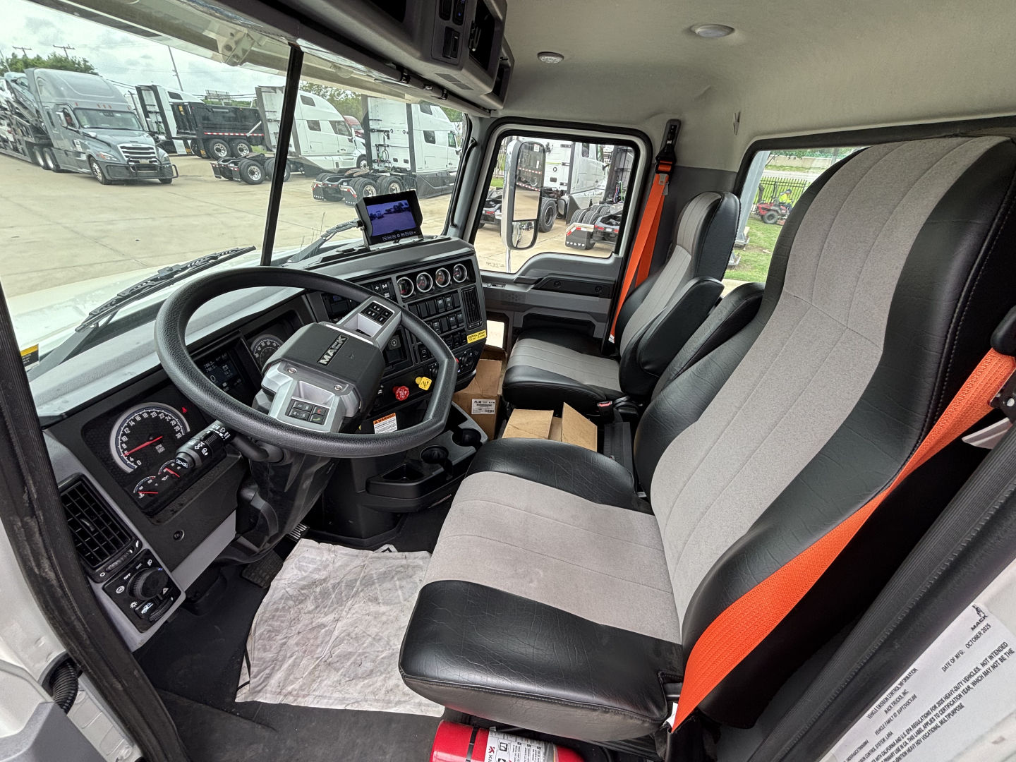 Interior of a Mack GR64F truck cab showing steering wheel, dashboard, and two-tone seats with orange seat belts.