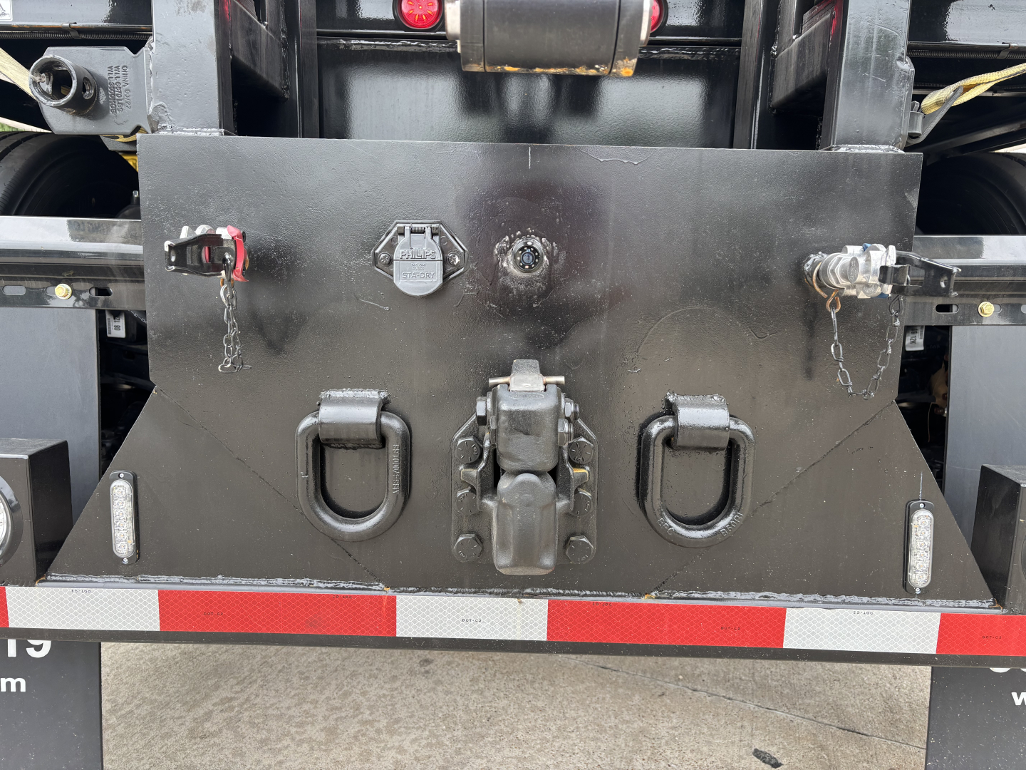 Crane truck rear platform showing two D-rings, central hitch, and control/valve panel with safety pins and chains.
