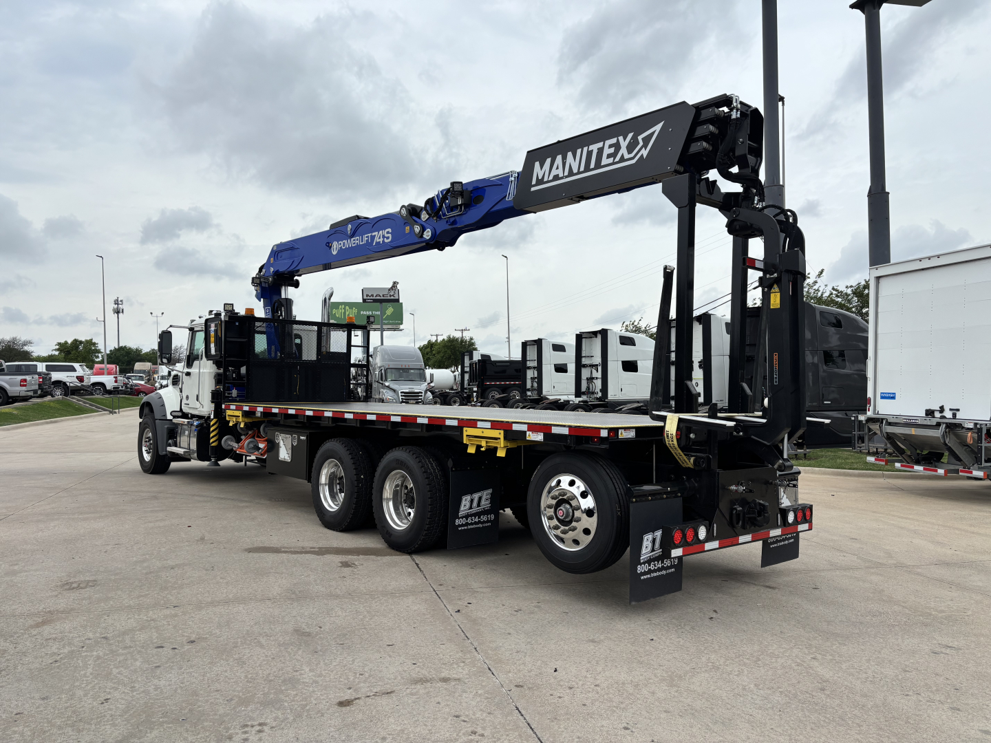 Truck-mounted crane on Mack GR64F with blue Manitex boom extending over flatbed trailer, rear stabilizers, and multiple axles in a dealership lot.