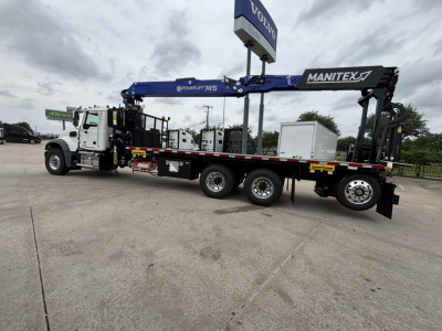 Mack GR64F crane truck with long blue Manitex boom mounted on a white cab and black trailer. Visible text includes MANITEX on the boom, POWERLIFT 7S, VOLVO sign overhead, and various labels along the crane.
