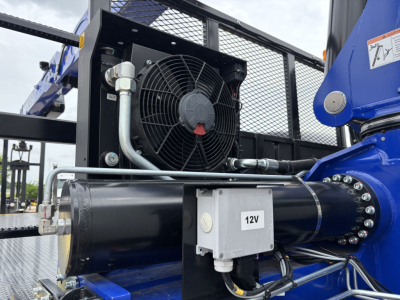 Crane truck engine bay area showing 12V auxiliary control box, radiator fan, hydraulic hose, and blue boom component on Mack GR64F.