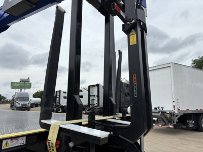 Mack GR64F crane truck components in view: hydraulic boom supports, black structural members, control rail, and warning label on upright; flatbed surface visible with other trucks in background.