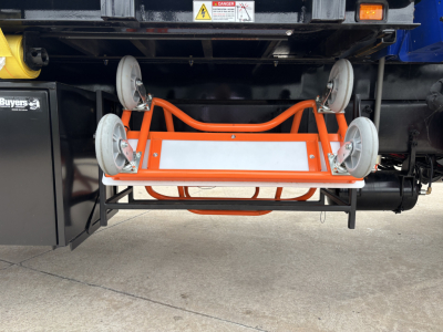 Mack GR64F crane truck underside with orange trolley and four caster wheels, visible warning labels, and hydraulic components.
