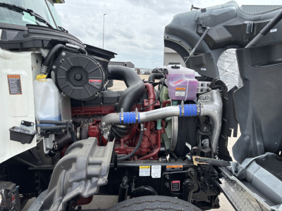 Mack GR64F engine bay with visible turbocharger, cooling reservoirs, hoses, and pink coolant tank. Labeling and components visible from driver side.
