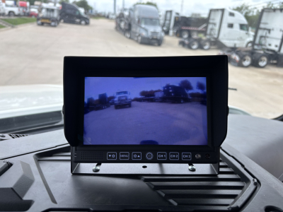 In-cab monitor screen with control panel showing multiple labeled buttons; ambient view of trucks in a lot through the screen.