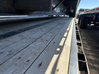 Flatbed trailer deck with steel planks and edge rails; visible are wooden deck boards, shadows, and part of a truck trailer frame.