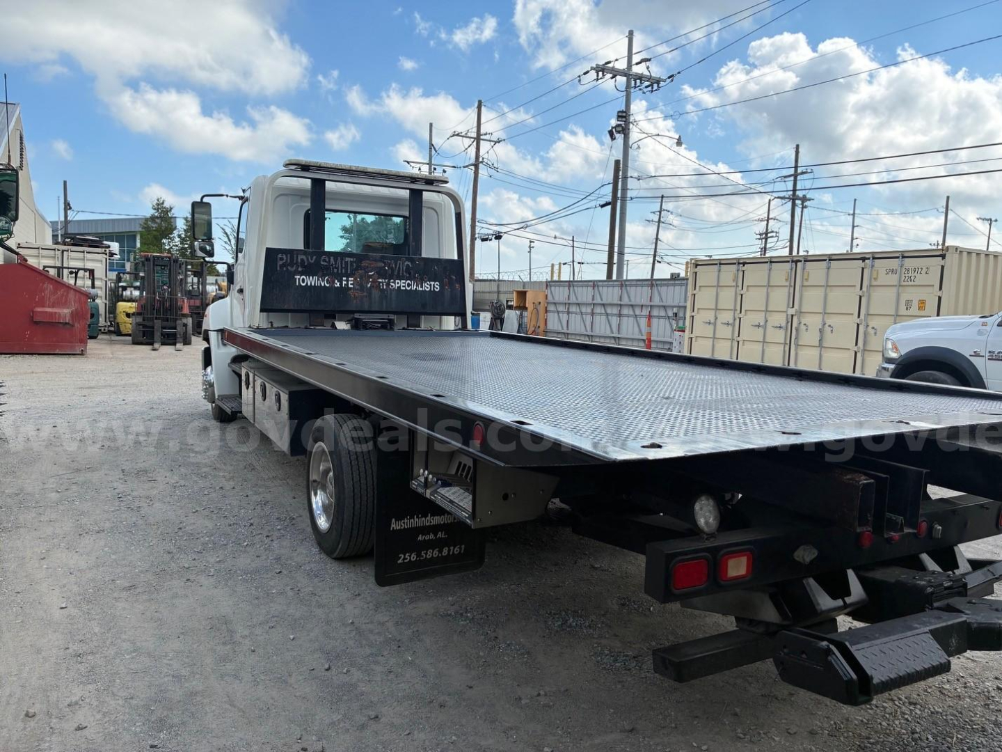 Flatbed tow truck with a long metal bed and white cab. On the rear tail, red lights and bumper. Side window has white text reading 'P..' and 'SPECIALISTS'. There are utility poles and storage containers in the background.