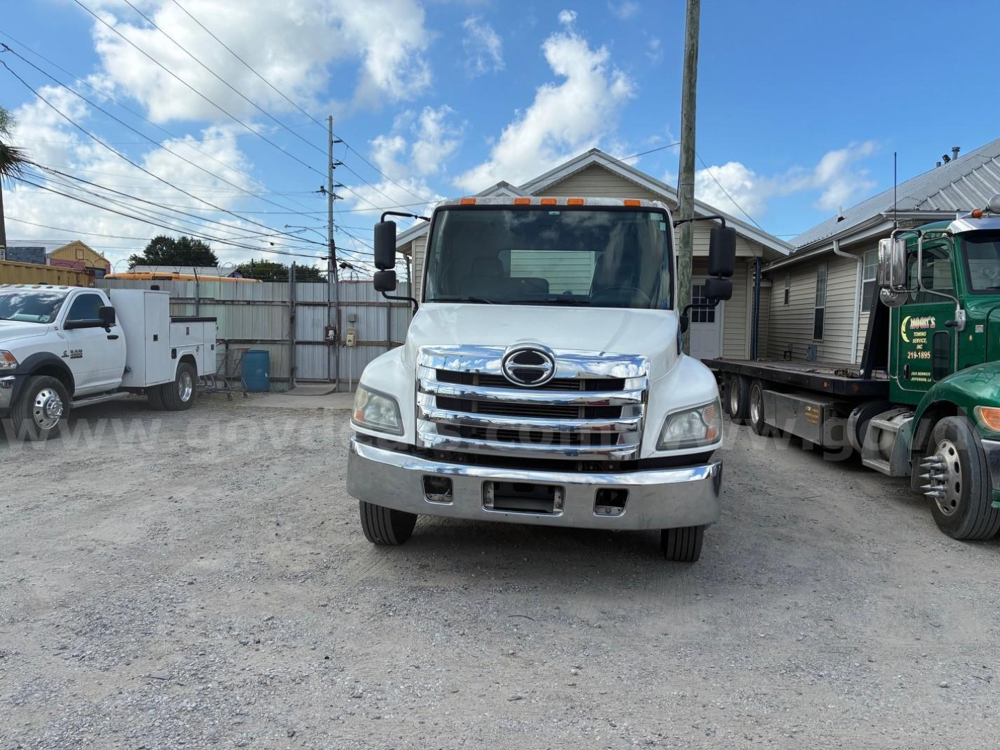 Front view of a white 2013 Hino conventional cab truck in a gravel lot, chrome grille, black mirrors, orange clearance lights on cab roof, other trucks in background.