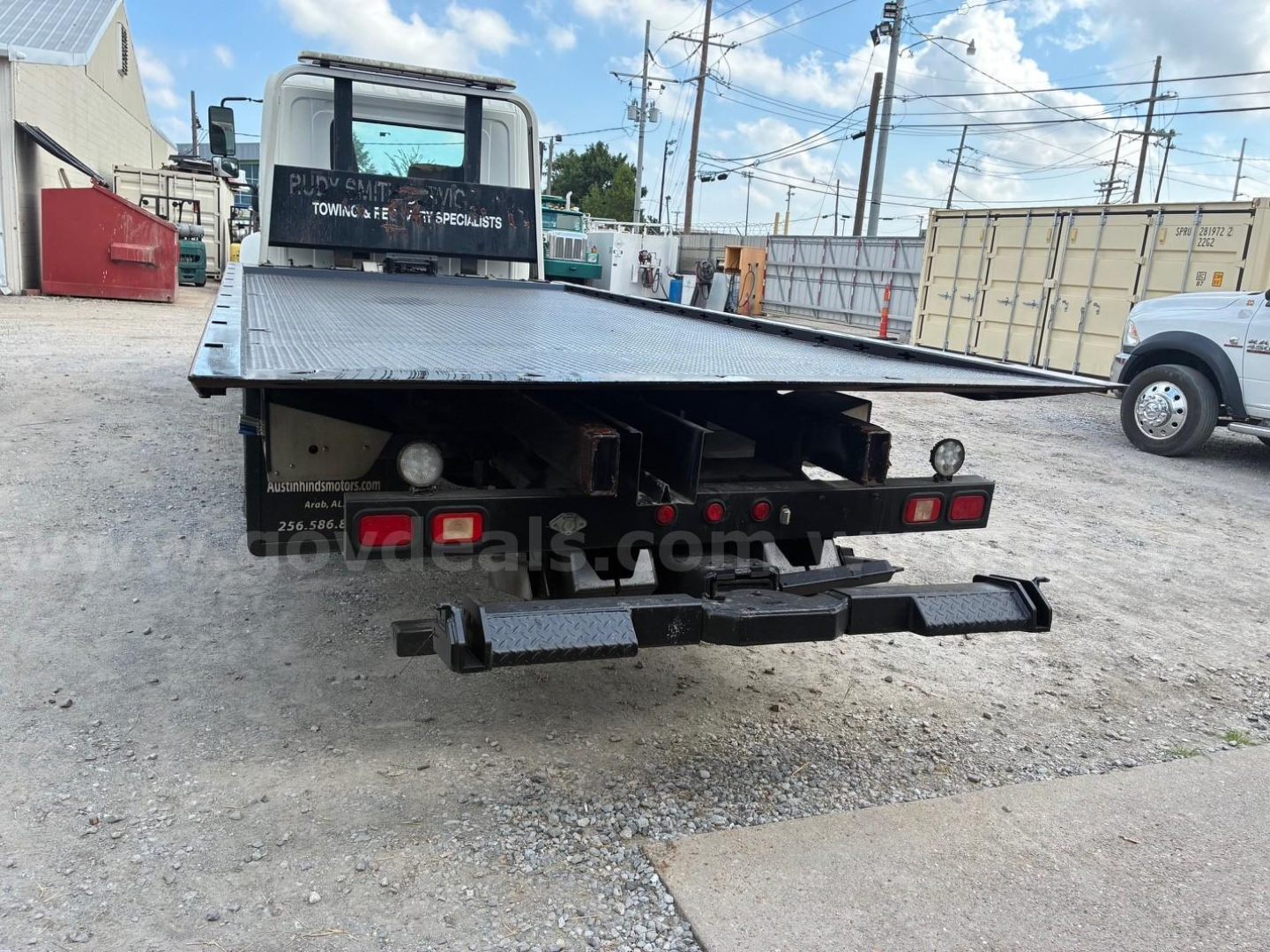 Flatbed tow truck rear view showing platform bed and bumper, Hino cab visible in background, gravel lot with shipping containers.